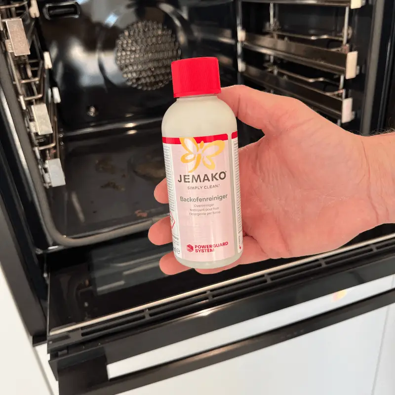 Eine Hand hält die Flasche des JEMAKO Backofenreinigers griffbereit vor einen geöffneten, leicht verschmutzten Backofen – JEMAKO Beratung und Verkauf in Steinhagen.